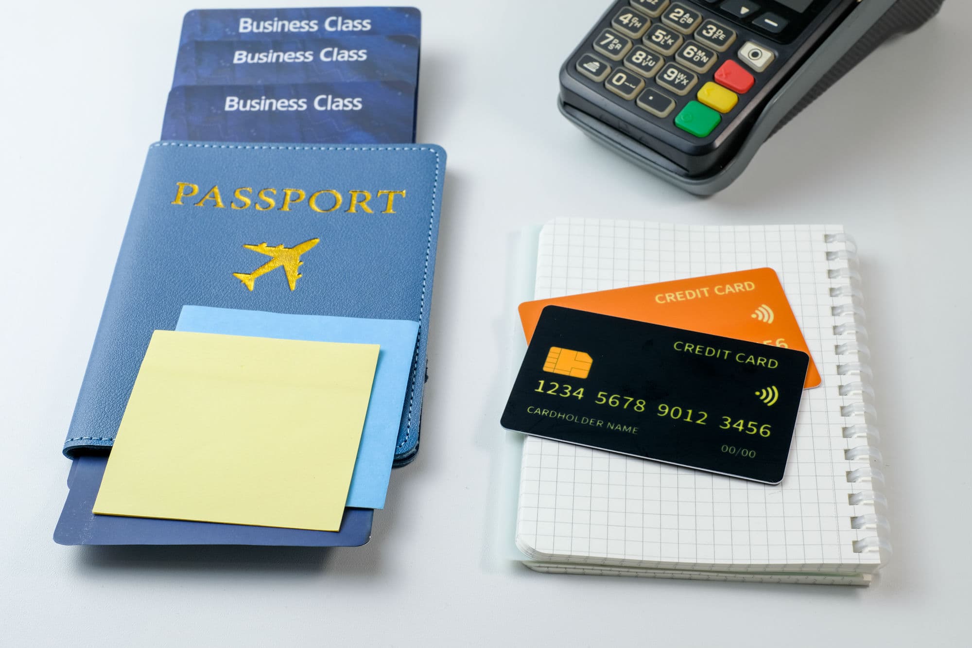 Un pasaporte y boletos de clase ejecutiva junto a dos tarjetas de crédito, una libreta y una terminal de pago sobre un escritorio blanco